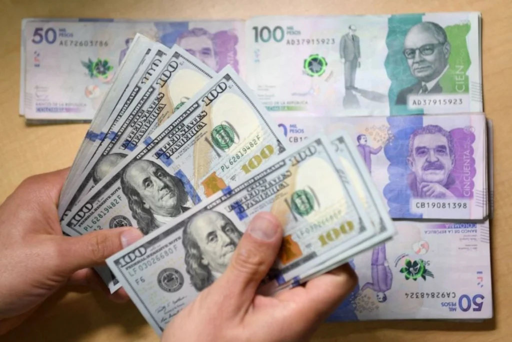 El dólar sigue bajando y estas son las claves para el bolsillo de los consumidores manos sosteniendo dólares con fondo de pesos colombianos sobre una mesa